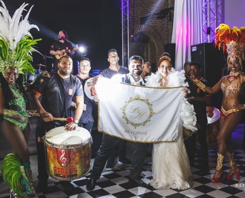 Casal Daiane e Heberson com equipe de bateria posando para foto junto com bandeira personalizada